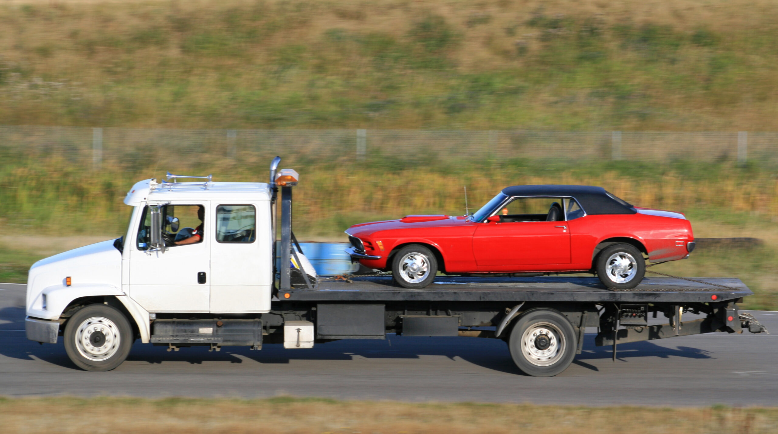 A red sports car being towed by a tow truck. Hauling, freight, or trucking theme. This image is a pan shot. Flat deck truck with automobile. Rural highway. White heavy duty truck with winch and loader. Flatbed.